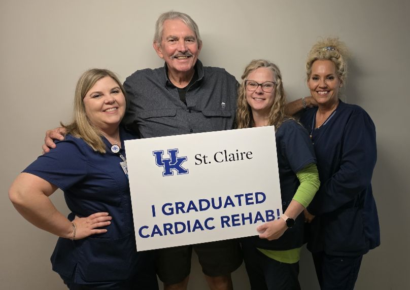 Three women in scrubs stand by a man in a gray button-down shirt and shorts. They're holding a sign that reads "UK St. Claire. I graduated Cardiac Rehab!"