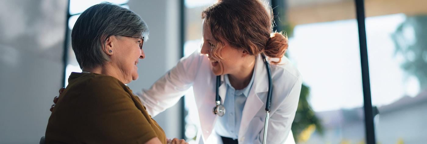 A female doctor wearing a white coat smiles as she speaks with and touches the shoulder of a female patient. The two are indoors, in an office with large, sunny windows.