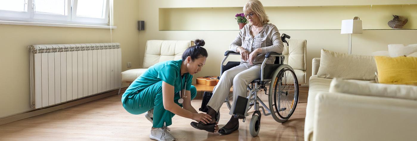 A woman wearing teal scrubs helps put a shoe on a woman who is seated in a wheelchair.